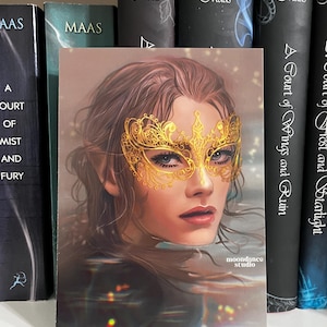 Puede incluir: Una impresión artística que muestra a una mujer con cabello castaño y una máscara dorada. La imagen está colocada frente a libros, incluido uno titulado "A Court of Wings and Ruin". El nombre del estudio del artista, "moondance studio", también es visible.