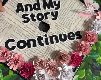 May include: A decorated graduation cap with the words "And My Story Continues" in bold black letters. The cap is adorned with pink, white, and silver paper flowers and a butterfly. The background is a page from a book.