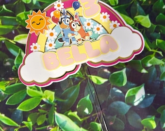Könnte beinhalten: Ein mehrschichtiger Tortenstecker mit den Zeichentrickfiguren Bluey und Bingo, einem Regenbogen, einer lächelnden Sonne und weißen Wolken. Der Aufsatz zeigt den Namen BELLA in großen rosa Buchstaben und die Zahl 3 auf einem transparenten Stab.