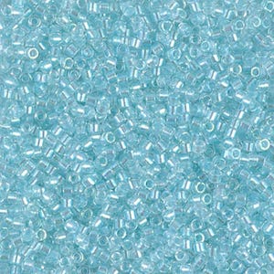 May include: A close-up shot of a pile of small, light blue seed beads. The beads are cylindrical with a hole running through the center, and they have a slightly iridescent quality. The beads are scattered, creating a textured surface.