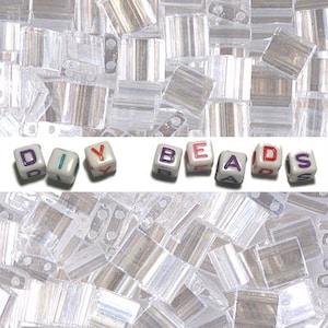 May include: DIY Beads spelling out the word "DIY BEADS" in white letter beads with purple and red accents. The beads are on a white background with a pile of clear glass beads in the background.