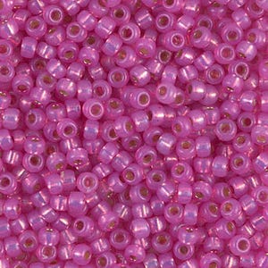 May include: Close-up of a pile of small, round, pink seed beads. Each bead has a small hole and a metallic gold-colored center. The beads are densely packed together, creating a textured surface.