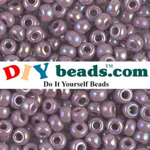 May include: A close-up image of a large quantity of small, iridescent purple glass beads. The beads are round with a hole in the center. The beads are arranged in a random pattern. DIYbeads.com Do It Yourself Beads.