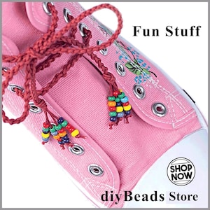 May include: A pink canvas sneaker featuring dark red braided laces adorned with multi-colored beads at the ends. The shoe includes silver metal eyelets and white stitching. Text on the image reads Fun Stuff and SHOP NOW diyBeads Store.
