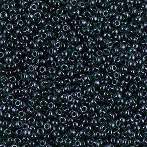 May include: A close-up view of a large quantity of dark green glass seed beads. The beads are small and round, with a hole in the center. They are arranged in a random pattern, creating a textured surface.