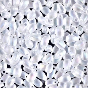 May include: A close-up of a pile of white iridescent glass beads. The beads are oval shaped with a hole at the top.