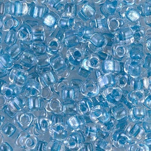 May include: A close-up of a large quantity of small, light blue, transparent glass beads with a shimmery finish. The beads are shaped like a rounded rectangle with a hole in the center.