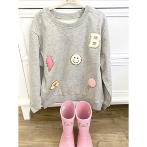 May include: A grey sweatshirt with a variety of colourful patches, including a lightning bolt, a smiley face, a rainbow, a balloon, and a letter "B". The sweatshirt is hanging on a white hanger. A pair of pink rain boots are in the foreground.