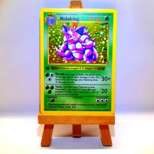 Könnte beinhalten: Eine holografische Nidoking-Sammelkarte mit einer lila Kreaturenillustration, 90 KP und Text zu den Attacken Thrash und Toxic. Die Karte zeigt einen Erstausgaben-Stempel, 140 cm und 62 kg, und steht auf einer kleinen hölzernen Staffelei.