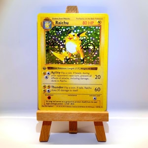 Könnte beinhalten: Eine Raichu-Sammelkarte mit einer gelben Figur mit blitzförmigem Schwanz, 80 KP und holografischen Sternenmustern. Die Karte zeigt die Angriffe Agility und Thunder sowie einen First-Edition-Stempel und steht auf einer kleinen Holzstaffelei.