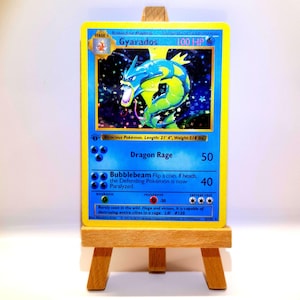 Könnte beinhalten: Eine holografische Gyarados-Sammelkarte mit blauem Rand, die ein blau-gelbes Seeschlangen-Motiv, 100 KP sowie die Attacken Drachenwut und Blubbstrahl zeigt. Der Kartentext enthält Atrocious Pokémon, Länge: 6,5 m und Gewicht: 235 kg.