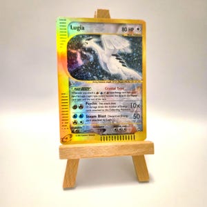 Może przedstawiać: Karta kolekcjonerska Lugia z 80 HP i umiejętnością Crystal Type umieszczona na małej drewnianej sztaludze. Karta zawiera ilustrację białego stworzenia, ataki Psychic i Steam Blast oraz tekst opisujący mechanikę gry na żółtym tle.