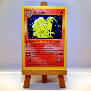 Könnte beinhalten: Eine holografische Ninetales-Sammelkarte mit einer gelben Fuchsgrafik, 80 KP sowie den Attacken Lure und Fire Blast. Die Karte zeigt ein 1st Edition-Symbol, eine Länge von 109 cm und ein Gewicht von 20 kg. Sie steht auf einer kleinen Holzstaffelei.