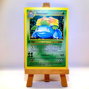 Könnte beinhalten: Eine holografische Venusaur-Sammelkarte mit einem blauen Wesen, das eine große Blume auf dem Rücken trägt, 160 KP und Text, der die Pokémon-Power Energy Trans und die Solarstrahl-Attacke beschreibt, auf einer kleinen Holzstaffelei.