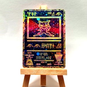 May include: A rectangular trading card with a holographic finish resting on a small wooden easel. The card features a stylized outline of a creature in a central frame against a colorful nebula background, surrounded by cryptic symbols and text.