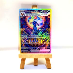 Könnte beinhalten: Eine Umbreon EX Sammelkarte mit einer lebendigen, mehrfarbigen holografischen Illustration des Charakters, umgeben von leuchtenden Elementarsymbolen, präsentiert auf einer kleinen Holzstaffelei. Der Kartentext enthält Moon Mirage.