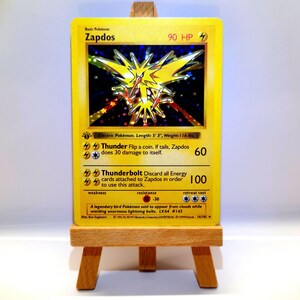 Könnte beinhalten: Eine gelbe, holografische Zapdos-Sammelkarte mit dem Elektro-Pokémon und 90 KP. Die Karte zeigt die Attacken Donner und Donnerblitz, ein 1st-Edition-Symbol sowie Textangaben zu einer Größe von 160 cm und einem Gewicht von 52,6 kg.