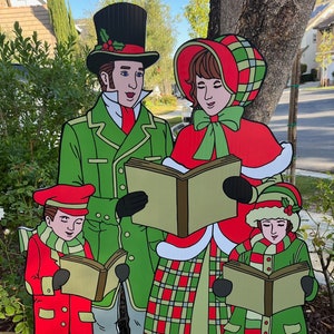 May include: A decorative outdoor Christmas carolers display. The family of four is dressed in festive attire, with green and red coats, hats, and scarves. They are holding songbooks and singing carols. The display is made of a durable material.