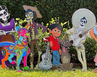 May include: A collection of colorful cardboard cutouts featuring characters from the animated movie "Coco." The characters include skeletons, a boy, a dog, and a decorated horse, all standing on green grass.