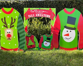 May include: A festive outdoor display featuring large, corrugated cardboard cutouts of ugly Christmas sweaters. Designs include a reindeer, snowman, and Christmas tree. A photo frame with the text "'tis the season to rock ugly sweaters" completes the scene.