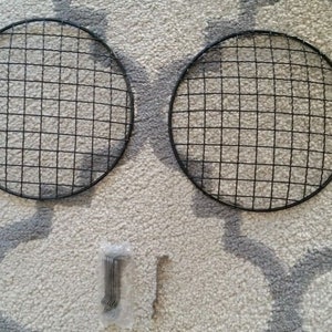 May include: Two round, black metal grill grates with a grid pattern, and a small package of metal fasteners. The grates are circular, with a diameter of approximately 10 inches. The items are on a patterned, light-colored rug.