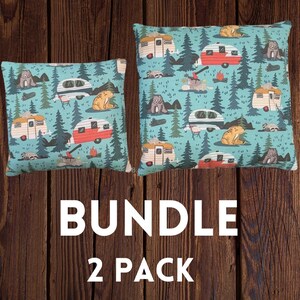 May include: Two turquoise blue throw pillows with a camping theme. The pillows feature a repeating pattern of vintage campers, bears, and trees. The text "BUNDLE 2 PACK" is printed below the pillows.