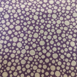 May include: Close-up of a fabric with a repeating pattern of white circles on a purple background. The circles vary in size and are densely packed, creating a textured appearance. The fabric has a woven texture.