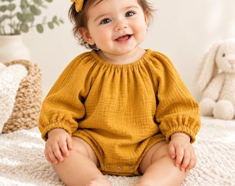 May include: A mustard yellow long-sleeved romper made of textured muslin fabric, featuring a gathered round neckline, elasticized cuffs, and metal snap closures at the bottom. A small matching mustard yellow bow hair clip is worn.