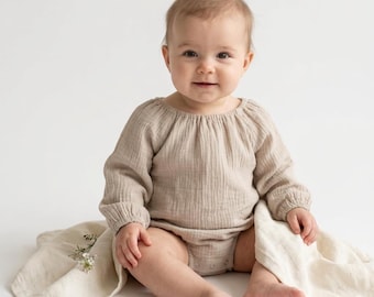 May include: A beige long-sleeved baby bodysuit made of textured muslin fabric with a gathered neckline and elasticized cuffs, worn by a baby sitting on a matching beige muslin cloth with a small sprig of white flowers.