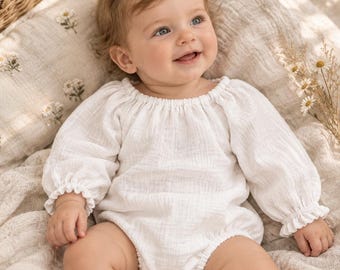 May include: A long-sleeved white baby romper made of textured muslin fabric. The garment features a gathered round neckline, elasticized cuffs at the wrists, and a snap-button closure at the bottom for easy access. The fabric has a soft, crinkled appearance.