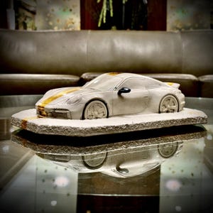 May include: A light gray sculpture of a sports car with gold racing stripes, mounted on a rectangular stone base. The car's design is detailed, and the base has a textured surface. The sculpture is reflected on a glass surface.