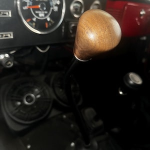 May include: Close-up of a wooden gear shift knob in a vehicle's interior. The knob is a light brown color and is attached to a black metal shift lever. A speedometer is visible in the background, along with other vehicle controls.