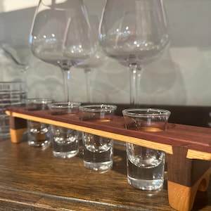 May include: A wooden shot glass holder with five clear shot glasses. The holder is made of dark red and light brown wood. Wine glasses are in the background.