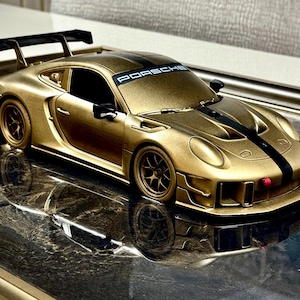 May include: A gold-colored toy sports car with black racing stripes and a rear spoiler. The word "PORSCHE" is displayed on the windshield. The car is on a reflective surface.