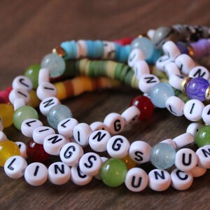 May include: A collection of colorful beaded bracelets. The bracelets feature white letter beads spelling out "LINGSUN" and other colorful beads. The bracelets are in shades of red, yellow, green, blue, and purple.