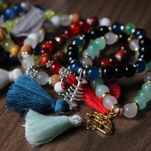 May include: A collection of beaded bracelets with colorful gemstones and tassels. The bracelets feature various colors, including red, blue, green, and black, with gold-colored charms and silver accents. The tassels are in shades of yellow, white, blue, and green.