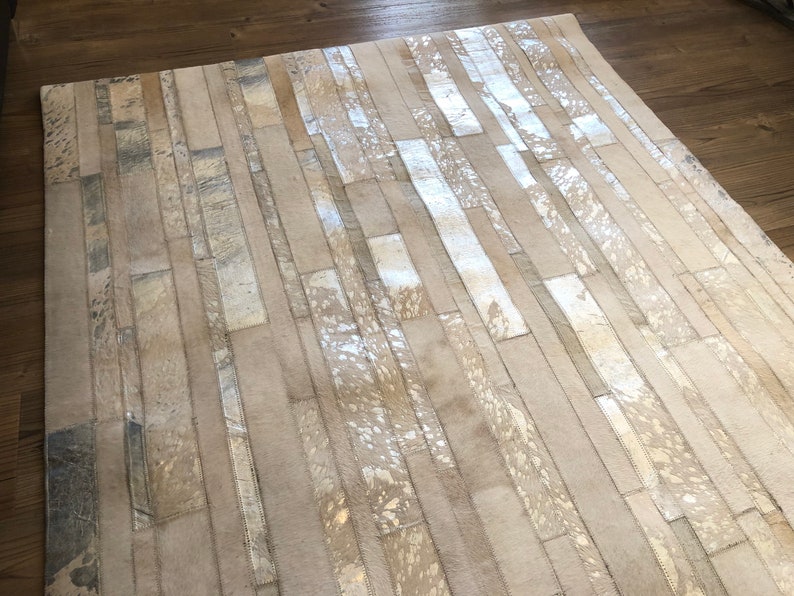 May include: A white and silver cowhide rug with a geometric pattern. The rug is made of strips of leather that are sewn together to create a unique design.