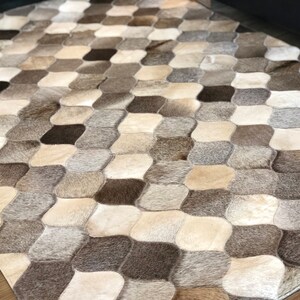 Könnte beinhalten: Ein Patchwork-Teppich aus Kuhfell in Braun-, Grau- und Weißtönen. Der Teppich besteht aus einzelnen Fellstücken, die zusammengenäht wurden, um ein geometrisches Muster zu bilden.
