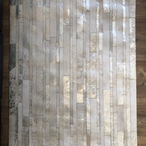 May include: A white and silver cowhide rug with a patchwork design. The rug is made of strips of leather that have been sewn together to create a unique pattern.