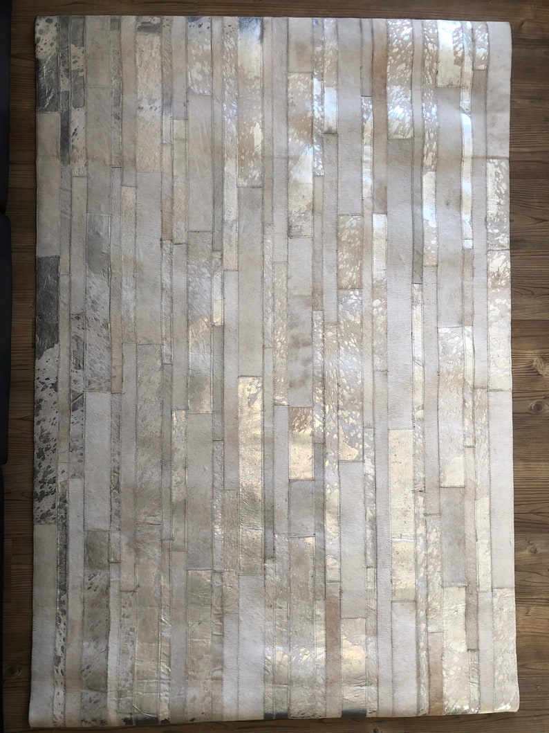 May include: A white and silver cowhide rug with a patchwork design. The rug is made of strips of leather that have been sewn together to create a unique pattern.