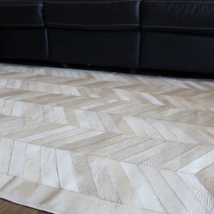 May include: A white and beige cowhide rug with a herringbone pattern. The rug is made of natural leather and has a rustic, farmhouse style.