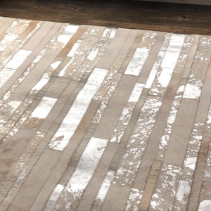 May include: A white and silver cowhide rug with a patchwork design. The rug is made of strips of leather that have been sewn together to create a unique pattern.