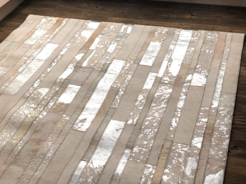 May include: A white and silver cowhide rug with a patchwork design. The rug is made of strips of leather that have been sewn together to create a unique pattern.