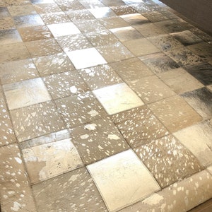 Puede incluir: Una alfombra de piel de vaca patchwork en un esquema de colores plateado y blanco. La alfombra está hecha de piezas individuales de piel de vaca que se han cosido juntas para crear un diseño único y elegante.