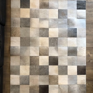 Puede incluir: Una alfombra rectangular construida a partir de una cuadrícula de parches cuadrados de piel de vaca. La alfombra presenta un patrón de tablero de ajedrez variado en tonos crema, beige, gris claro, carbón y marrón oscuro con texturas naturales.