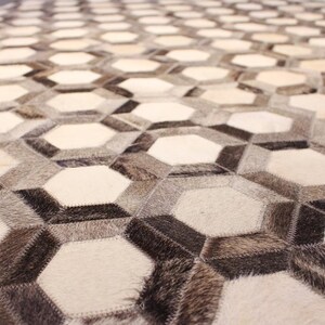 May include: Close-up of a rug with a geometric pattern. The rug has a repeating design of interconnected hexagons in white, grey, and brown. The texture appears to be a mix of smooth and slightly raised areas.