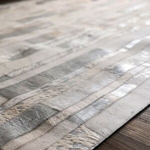 May include: A white and silver cowhide rug with a striped pattern. The rug is made of different pieces of cowhide that have been sewn together.