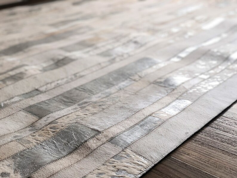 May include: A white and silver cowhide rug with a striped pattern. The rug is made of different pieces of cowhide that have been sewn together.