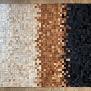 Może przedstawiać: Prostokątny dywan wykonany z kwadratowych kawałków skóry w odcieniach bieli, brązu i czerni. Dywan ma wzór gradientowy, przechodzący od bieli przez brąz do czerni.