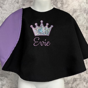 May include: Black cape with a lilac sleeve. The cape features a silver glitter crown and the name "Evie" embroidered in purple. The cape is displayed on a mannequin.